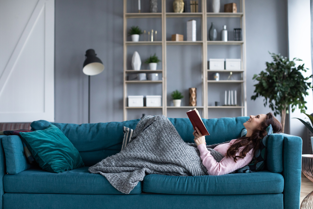 Braunhaarige lächelnde Frau, die in eine Decke gehüllt mit einem Buch auf dem Sofa liegt