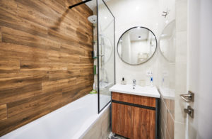 Stilvolle und komfortable Version des Badezimmerdesigns auf kleinem Raum. Weiße Farbe der rechteckigen Badewanne und des Waschbeckens in harmonischer Kombination mit rundem Spiegel, ergänzt durch Holzdekor.