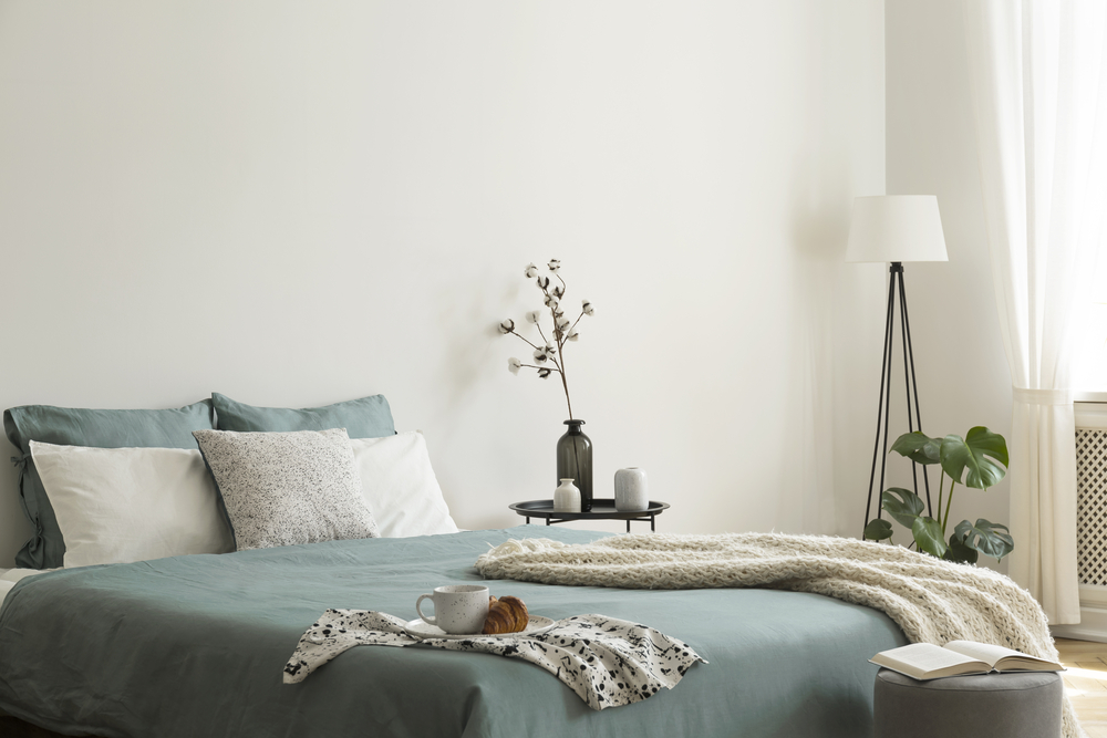 Schlafzimmereinrichtung mit salbeigrünen und weißen Laken und Kissen und einer Decke. Schwarzer Metalltisch mit Vasen neben dem Bett.