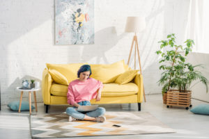 Junge Frau sitzt mit Laptop auf dem Boden vor einem gelben Sofa